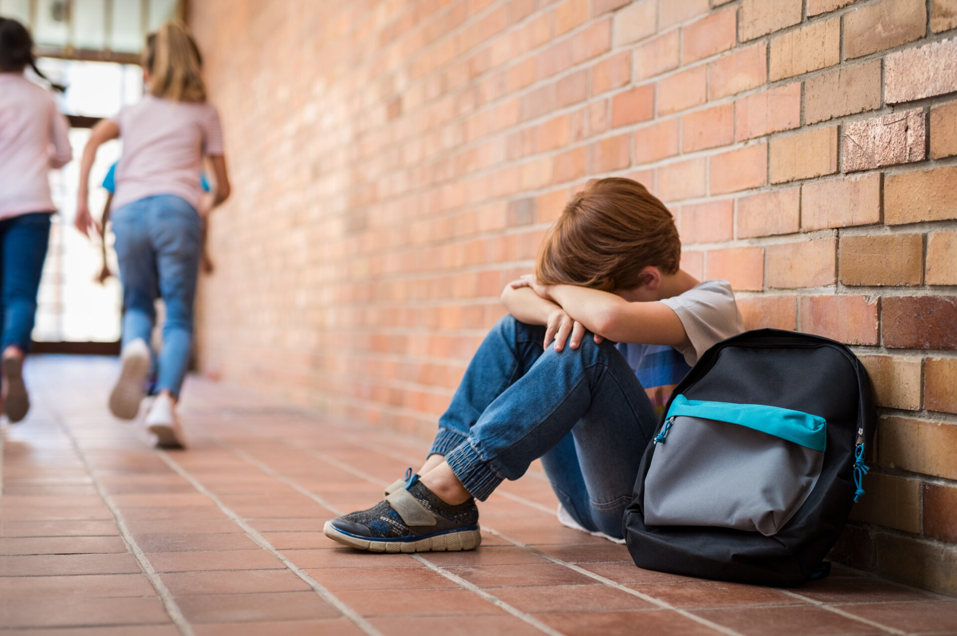 einen kleinen Jungen, der traurig und allein auf einem Schulflur sitzt, während andere Schülerinnen von ihm wegrennen